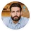 Portrait of a man with dark hair and beard wearing a light blue shirt against a plain background.