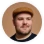 Man with a beard wearing a brown cap and a dark shirt, looking slightly to the side against a white background.